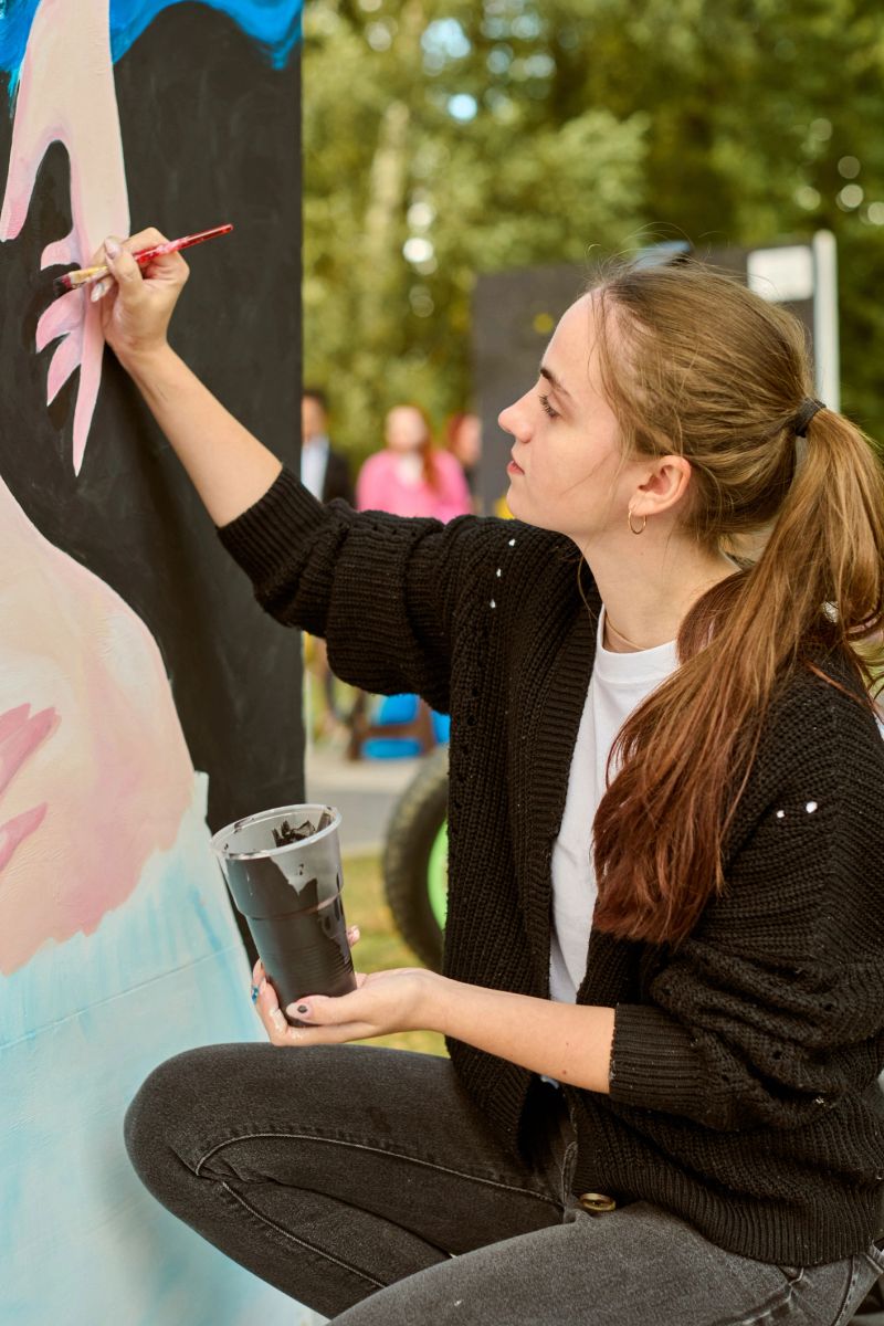 Young female painting outdoor mural focused expression brown hair casual attire. Art creation en plein air with natural light highlighting vibrant colors. Lush greenery surrounds