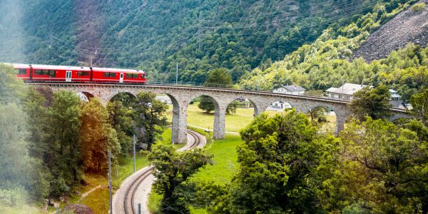 Red train crossing a stone viaduct over lush green landscape, glacier express train switzerland packages