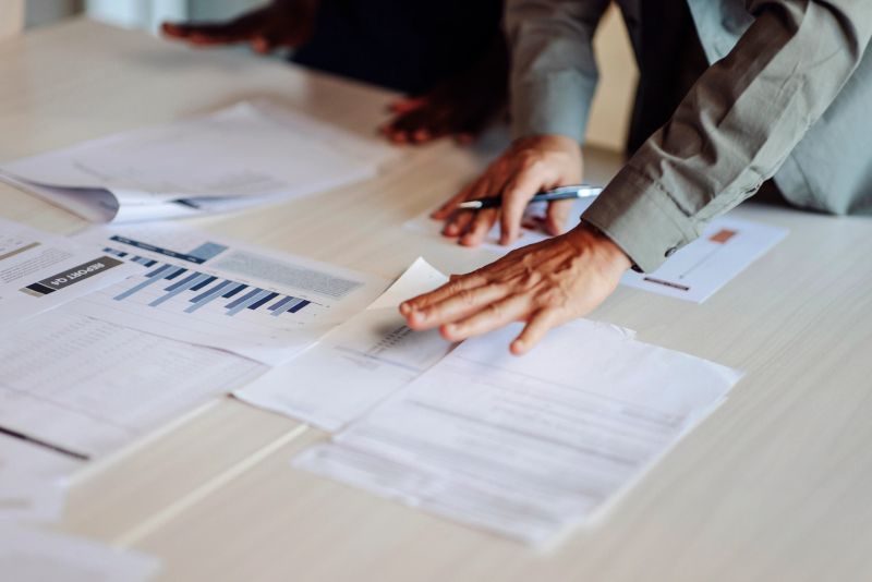 Professional teamwork showcasing colleagues reviewing financial data, graphs, and documents on a desk; Represents discussion, collaboration, and productivity in achieving business goals