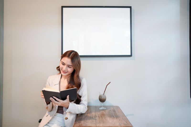 A woman is reading a book at a table. She is smiling and she is enjoying her time. a picture frame behind her.