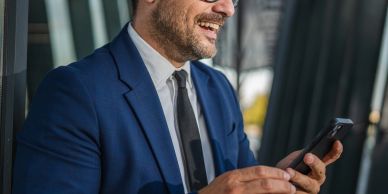 Smiling businessman in blue suit checking smartphone outside.