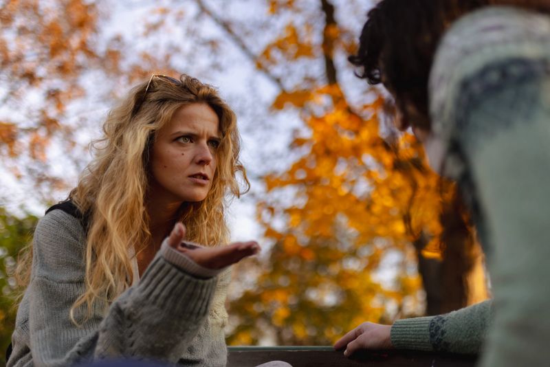 Two women appear deep in discussion, surrounded by vivid autumn leaves. One woman gestures expressively while the other listens intently. The warm light enhances the atmosphere.