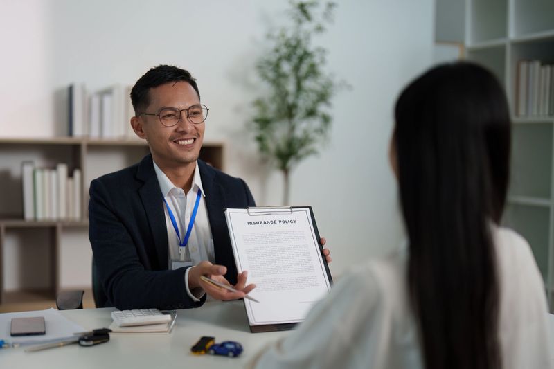 A smiling insurance agent presents a policy document to a client, highlighting key details in a bright office environment.