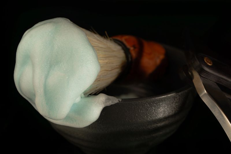 Shaving brush with teal lathered foam and straight razor in a dark bowl for men's grooming