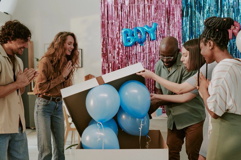 Group of young adult and middle aged multiethnic men and women opening large box with blue balloons at gender reveal party, smiling and clapping while celebrating together indoors