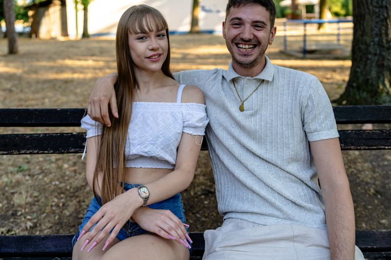 A cheerful duo enjoys their time in a serene park setting. They are seated together on a bench, sharing a moment of closeness and laughter. The natural sunlight enhances the vibrant and relaxed ambiance of the image, highlighting a friendly and joyful atmosphere.