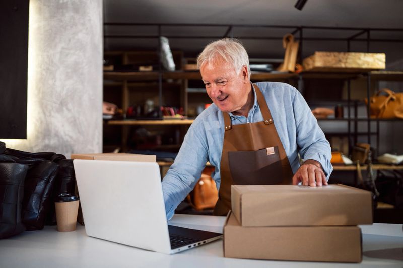 A senior craftsman working in a leather workshop, handling online orders, and packaging handmade products. Demonstrates dedication to craft and modern business practices.
