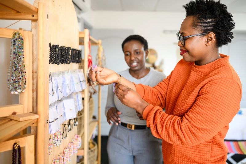 Two women enjoying shopping experience as one tests jewellery piece, inside creative retail studio filled with artisan crafts and handmade accessories.