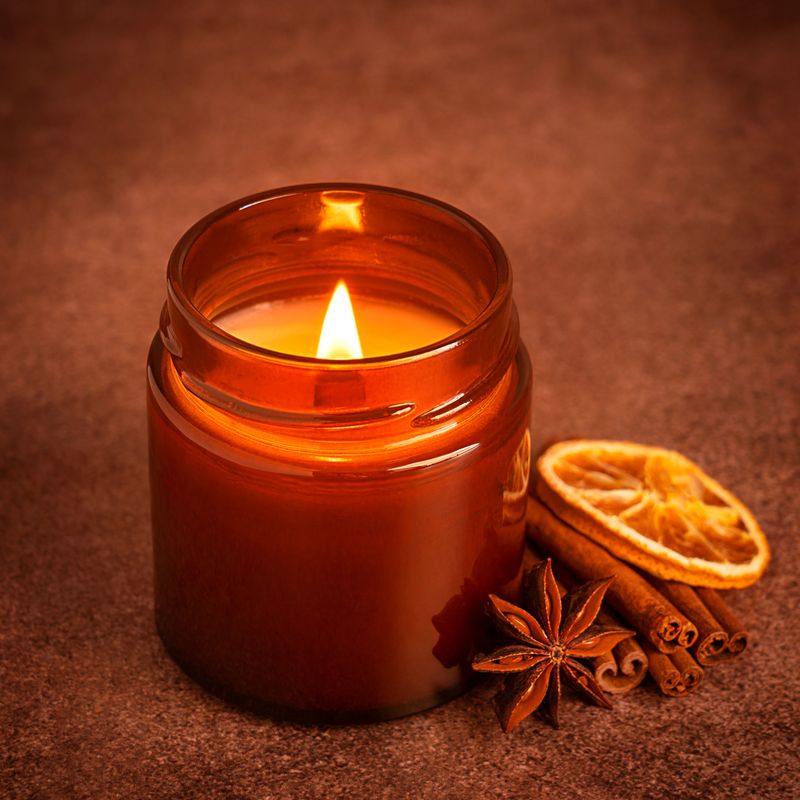 Cozy autumn glowing aromatic candle light in glass jar with cinnamon, star anise spice and dried orange fruit slice on dark brown rustic table prepared for evening winter aromatherapy or relaxation
