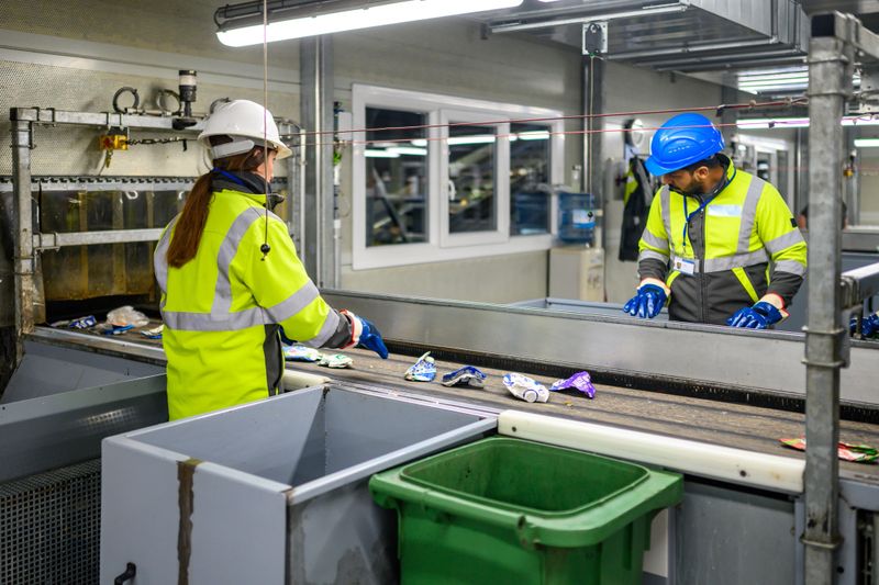A team of professionals in safety gear is overseeing the processing of materials on a conveyor belt in a waste management facility. They are sorting and managing recyclable items efficiently, featuring industrial equipment and organized operations.