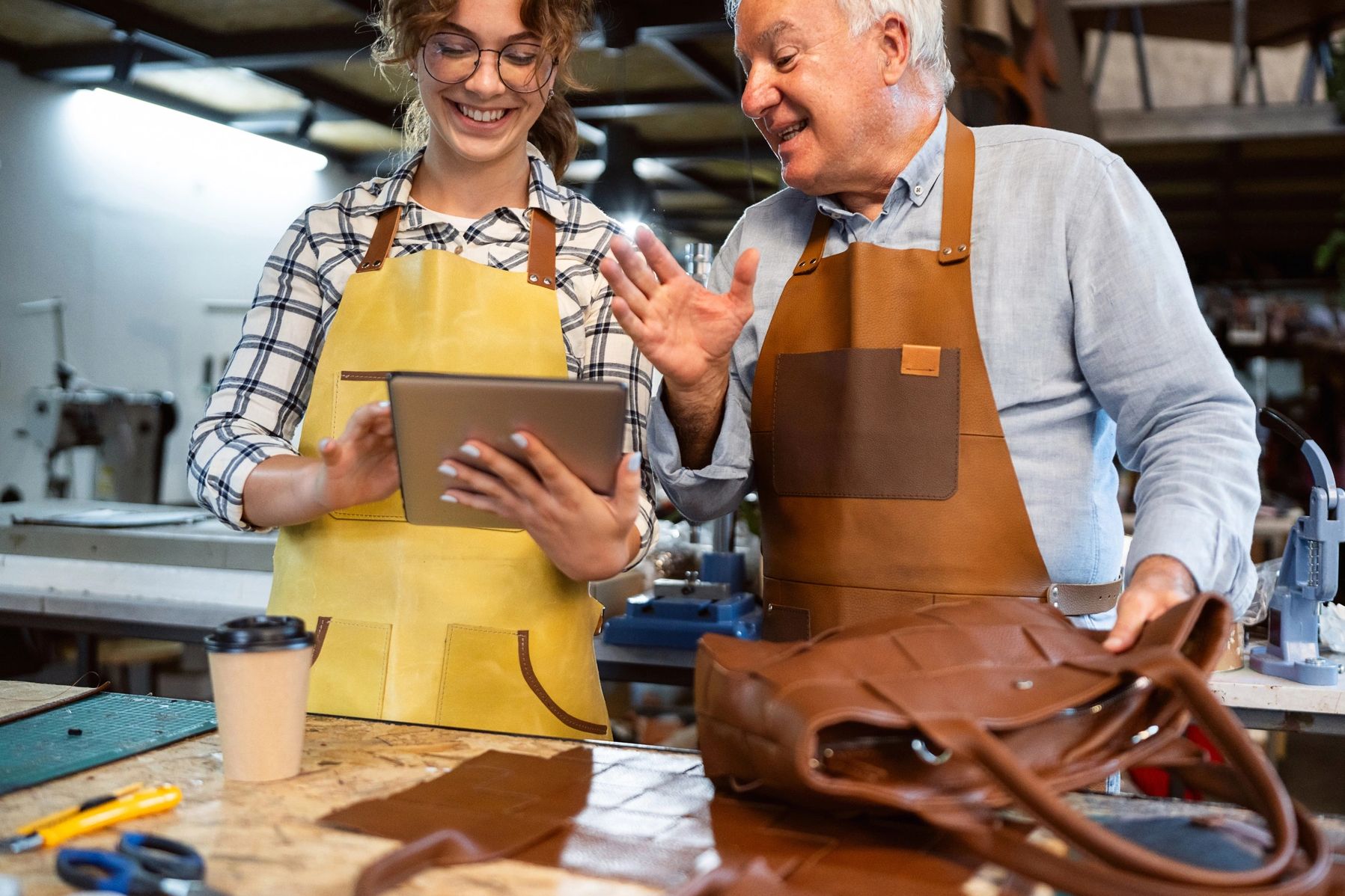 Two leather artisans smiling and discussing work using a tablet in a workshop.