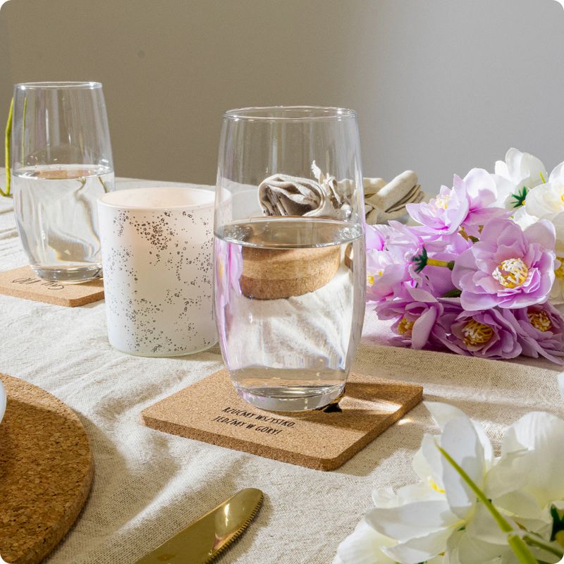 Closeup of a clear glass with water placed on a cork coaster on a linen table with flowers and soft sunlight.