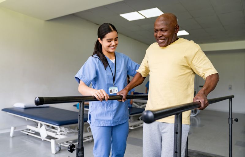 Happy physical therapist helping an African American senior man walking on bars at a rehabilitation center