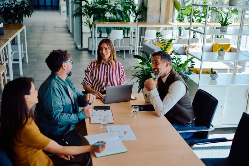 Group of adults engaged in a positive business meeting, discussing ideas with laptops and documents in a bright modern office filled with natural light and greenery