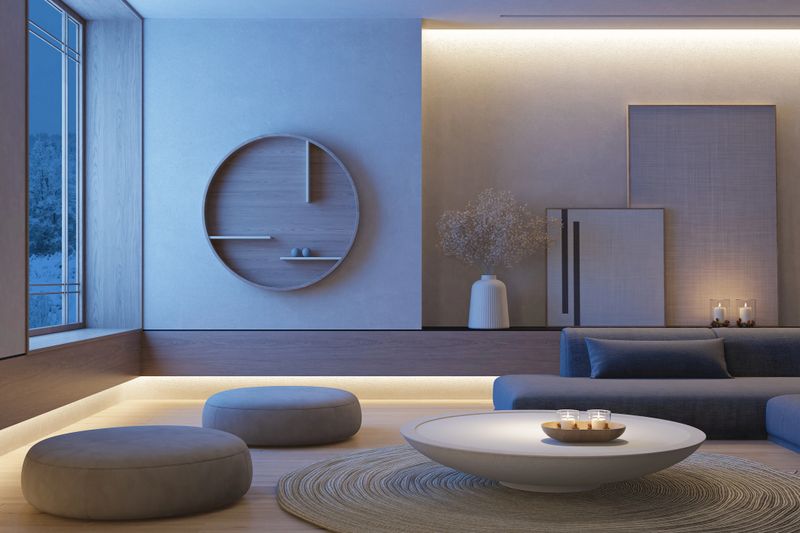 Night scene dusk blue hour. Living room with Japandi interior design, combining Japanese serenity with Scandinavian simplicity. The living room features a muted palette of gray, beige, and light wood tones, creating a tranquil and balanced atmosphere. Matte textures dominate the space—from the soft fabric of the sectional sofa to the natural wood flooring and understated decor. A round white coffee table and circular rug anchor the room with gentle symmetry. Minimalist accents like the dried floral arrangement, circular wall shelf, and leaning panels add visual interest without overwhelming the space. Natural light pours in through a large wooden-framed window, enhancing the organic feel of the materials. The modern ceiling light fixture complements the room’s clean lines and subtle geometry. This image is ideal for use in interior design portfolios, lifestyle blogs, furniture catalogs, or wellness-focused branding. It evokes a sense of calm, intentional living, and timeless style rooted in nature and simplicity.