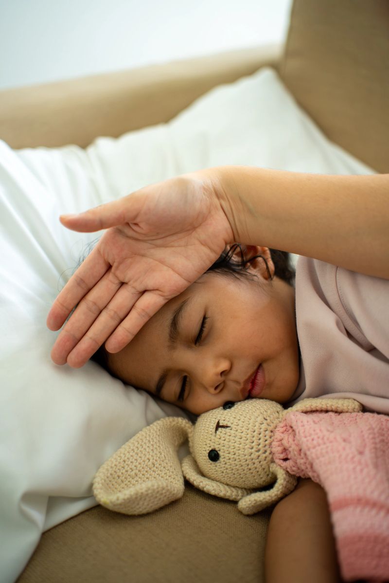 Vertical shot asian mother hand checking temperature of sick child’s fever on forehead while young daughter lays in bed, parent caring for child health insurance support and family medical coverage.