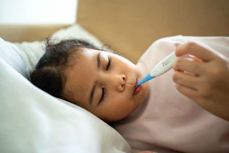 Mother using digital thermometer measuring body temperature of sick child’s fever lying on bed at home, parent caring for child health insurance support and family medical coverage.