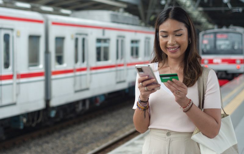 Image of an Asian woman using a credit card and smartphone, simulating an online ticket purchase while traveling. The background features a train and train platform, emphasizing modern technology in public transportation. Perfect for concepts of digital payment, online booking, smart travel, fintech, transportation lifestyle, and seamless commuting in urban environments.