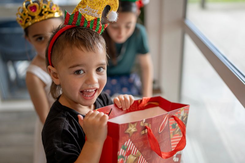 A young child wearing festive headbands smiles while holding a Christmas gift bag at home