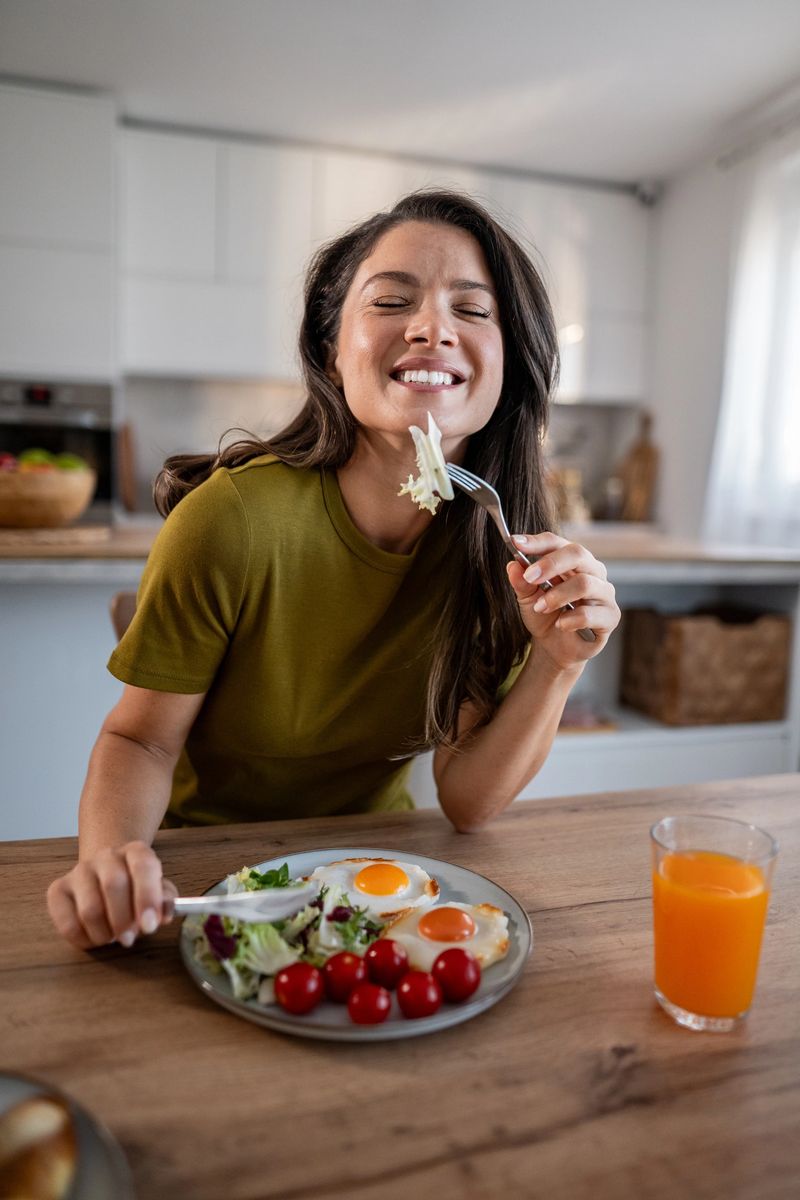 Woman closing eyes and smiling, savoring a healthy breakfast plate of fried eggs, salad, and cherry tomatoes, with orange juice on a wooden table in a well-lit kitchen