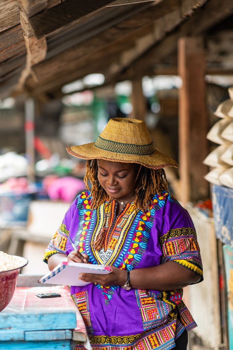 Small business owner, SME. Smiling traditional African market woman in a purple dashiki and straw hat takes inventory of her stock with a pen and notebook in front of her stall.