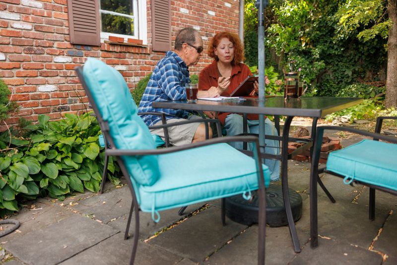 Female advisor with red hair meets with elderly man wearing glasses on outdoor patio. The man appears to have visual impairment as he reviews documents closely. They sit at dark metal table with turquoise cushioned chairs during daytime consultation. Brick house with white shuttered windows and lush garden plants create residential backdrop. Natural lighting illuminates the professional meeting space.