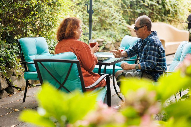 Insurance claims adjuster with red hair sits across from elderly man with visual impairment at outdoor patio table. The 47-year-old woman reviews paperwork and policy documents with the 82-year-old client during daytime consultation. Garden setting with lush green foliage surrounds the residential patio meeting space. Natural lighting illuminates the professional discussion between agent and policyholder. Selective focus on the consultation with soft background blur of garden plants.