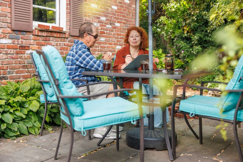 Insurance advisor with red hair meets with 82-year-old man who has vision impairment on outdoor patio. The woman reviews documents and coverage options while sitting at glass table with blue cushioned chairs. The senior man listens carefully during the consultation. Brick house with brown shutters and lush green landscaping provides backdrop for this home-based business meeting. Natural sunlight filters through trees during daytime session. Selective focus on the two people with garden elements softly blurred in foreground.