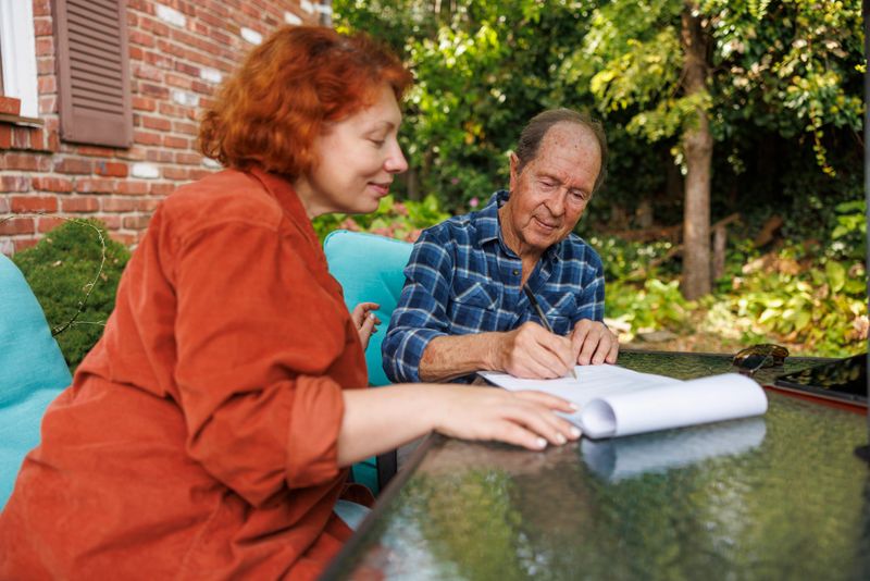 Mid-adult redhead woman in orange blazer sits beside elderly man with visual impairment at glass patio table. The woman guides the senior man through document signing process while he writes with pen on white papers. Meeting takes place on residential outdoor patio with brick house and mature landscaping visible in background. Natural daylight illuminates the scene during daytime consultation session. Close-up view captures collaborative interaction between service provider and client.