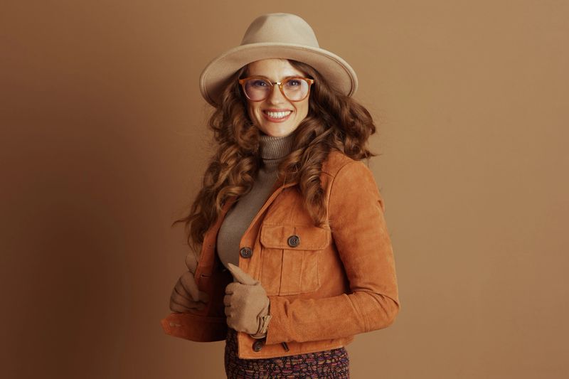 A fashionable woman with curly brown hair, wearing a tan fedora, orange suede jacket, and gloves, smiles while confidently adjusting her jacket against a warm brown background.