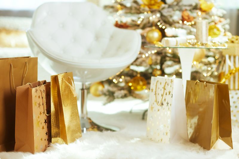 A luxurious collection of gold and brown gift bags rests on a plush white rug in front of a decorated Christmas tree and white armchair. The bright, festive scene creates a holiday atmosphere.