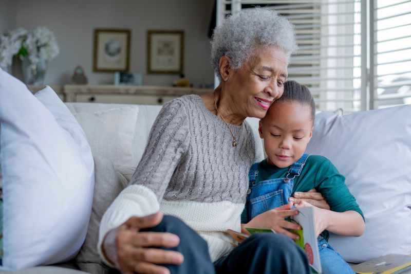 A warm, multigenerational moment as an elderly African American grandmother reads with her young grandchild on a comfortable living room sofa, highlighting family, love, and home comfort.