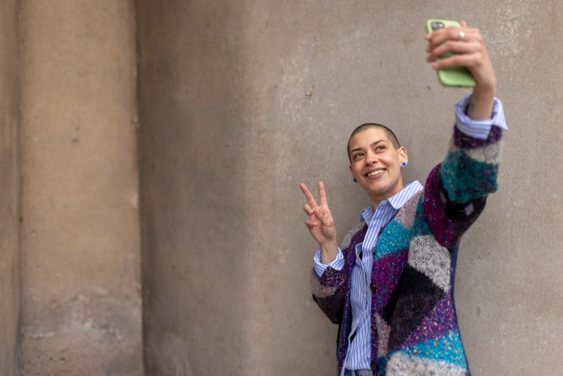 A cheerful bald woman in a vibrant patchwork jacket takes a selfie with a peace sign, standing against a neutral wall. Her confident smile radiates positivity and individuality.