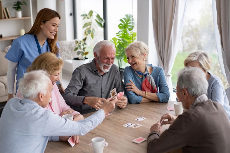 A friendly nurse assists a group of seniors enjoying a lively card game, filled with laughter and connection in a bright, welcoming setting.