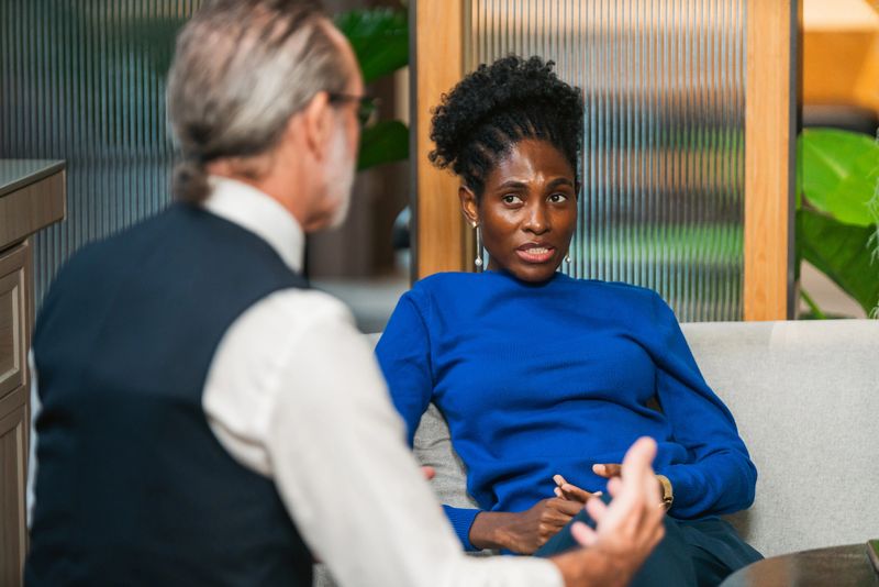 Two professionals converse on a sofa in a modern office lounge, engaged in a focused business discussion and mentoring session, conveying teamwork and communication in a casual workplace setting.