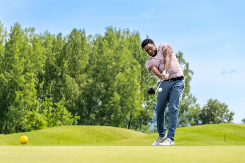 African American golfer in mid-swing on a lush green course, showcasing focus and athleticism. The vibrant setting highlights the sport's elegance, with a yellow ball in motion, emphasizing precision and skill in golf. Perfect for action-oriented sports imagery., captured with a medium shot.