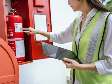 A woman in a safety vest inspecting a fire extinguisher with a tablet in hand.