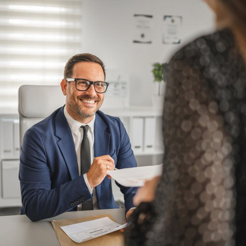 Professional businessman, wearing a suit and glasses, smiling while passing a document to a female colleague, sitting at his spacious office desk with a laptop and paperwork