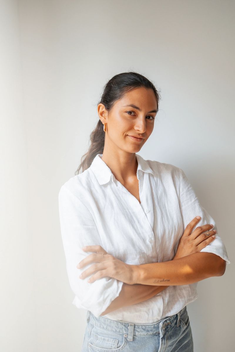 A confident young woman in casual attire standing against a white background while smiling. The image captures her relaxed and positive demeanor, illustrating themes of confidence, self-assurance, and natural beauty.