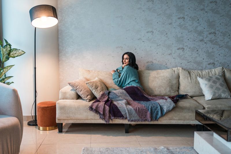 A young Arabic woman enjoys a tranquil moment on a plush sofa wrapped in a colorful blanket in a stylish, modern living room with soft lighting and elegant decor.
