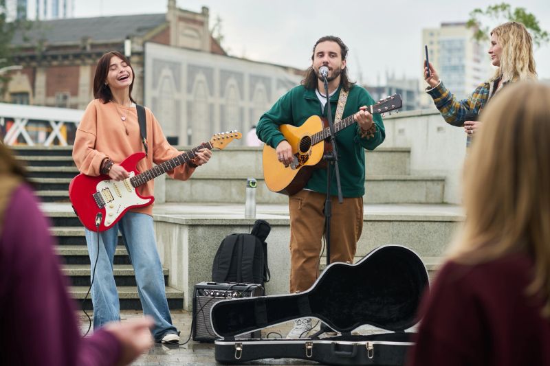 Young Caucasian woman playing electric guitar and singing with man playing acoustic guitar and singing outdoors while another woman recording performance on smartphone