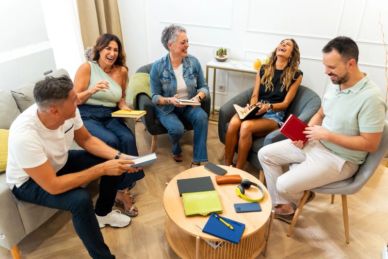 Group of five mixed-age range adults gathering in a living room, holding books and laughing, enjoying a book club meeting