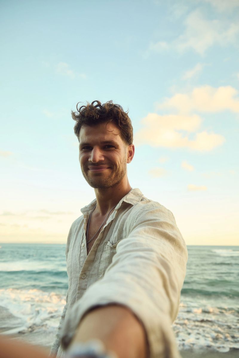 A smiling man in a light linen shirt captures a selfie at the seaside. Warm sunset light, calm sea, and clear sky create a relaxed, carefree mood ideal for travel and lifestyle themes.