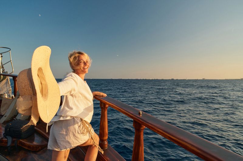 Woman on a boat holding a hat and smiling during sunset at sea.