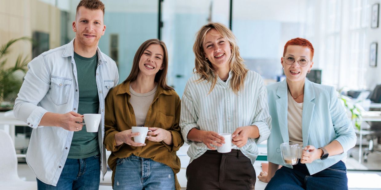 A group of four young adults smiling indoors with coffee mugs.