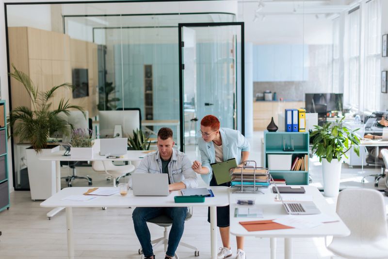 A male employee sitting at his desk working on a laptop, while a female colleague leans on the desk holding a folder. They are engaged in conversation, collaborating and solving a problem together in a modern office environment.