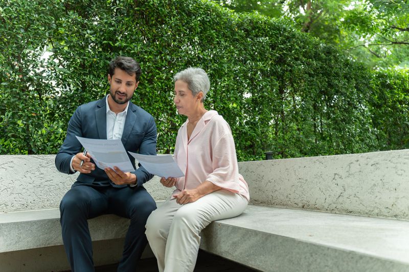 A Latin male Medicare insurance broker consults with an Asian senior woman outdoors, seated together on a stone bench surrounded by greenery. Both hold documents as the broker explains healthcare plan details while the senior carefully reviews the information. The candid and approachable moment conveys professionalism, trust, and personalized support, illustrating real-life Medicare enrollment and benefits planning in a relaxed outdoor setting