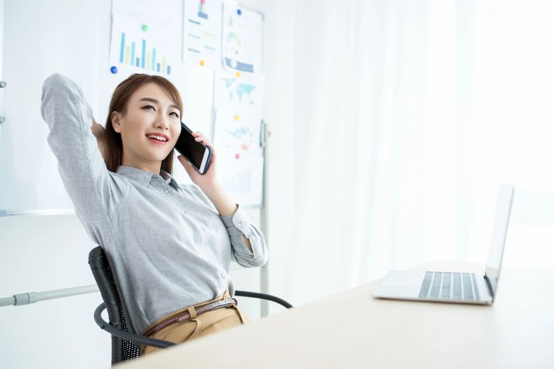 A woman is sitting in an office, talking on a phone with a relaxed posture, with a laptop on the desk and charts on the board in the background