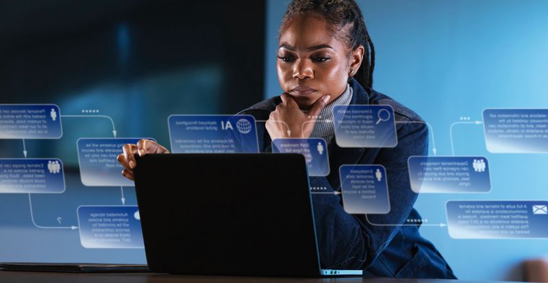 An African-American businesswoman concentrates as she uses her laptop in an office. A graphical overlay shows she is having an online conversation with an AI chatbot.