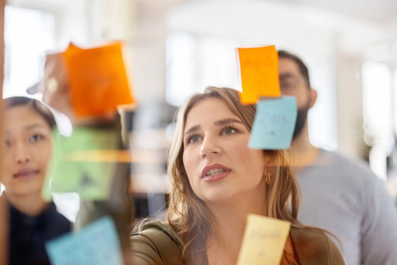 Confident female entrepreneur points to adhesive notes on a glass wall while discussing strategy with colleagues, showcasing agile methodology, innovation, and inclusive teamwork in a contemporary coworking space.